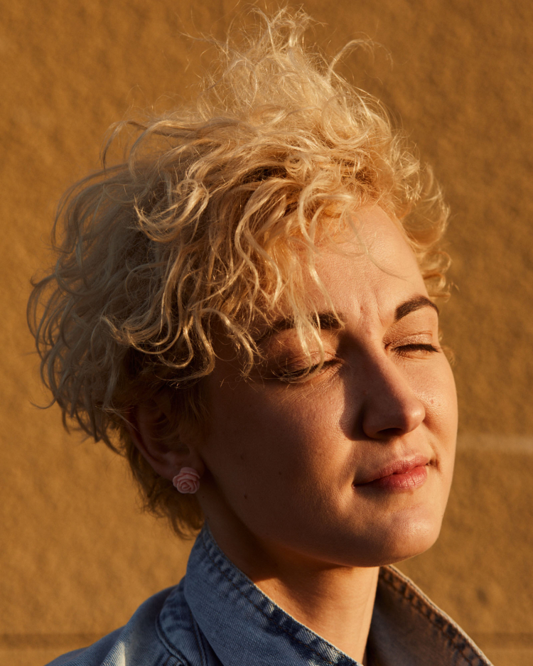 Close-up portrait of an adult woman with curly blonde hair, rose earrings, and a blue denim jacket, against a textured yellow background.