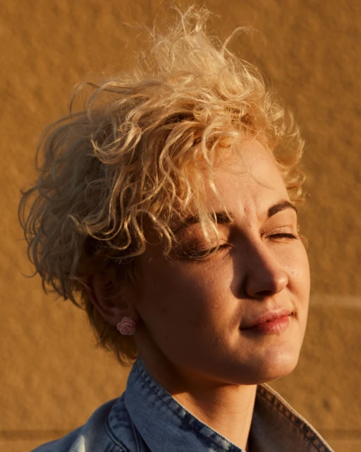 Close-up portrait of an adult woman with curly blonde hair, rose earrings, and a blue denim jacket, against a textured yellow background.