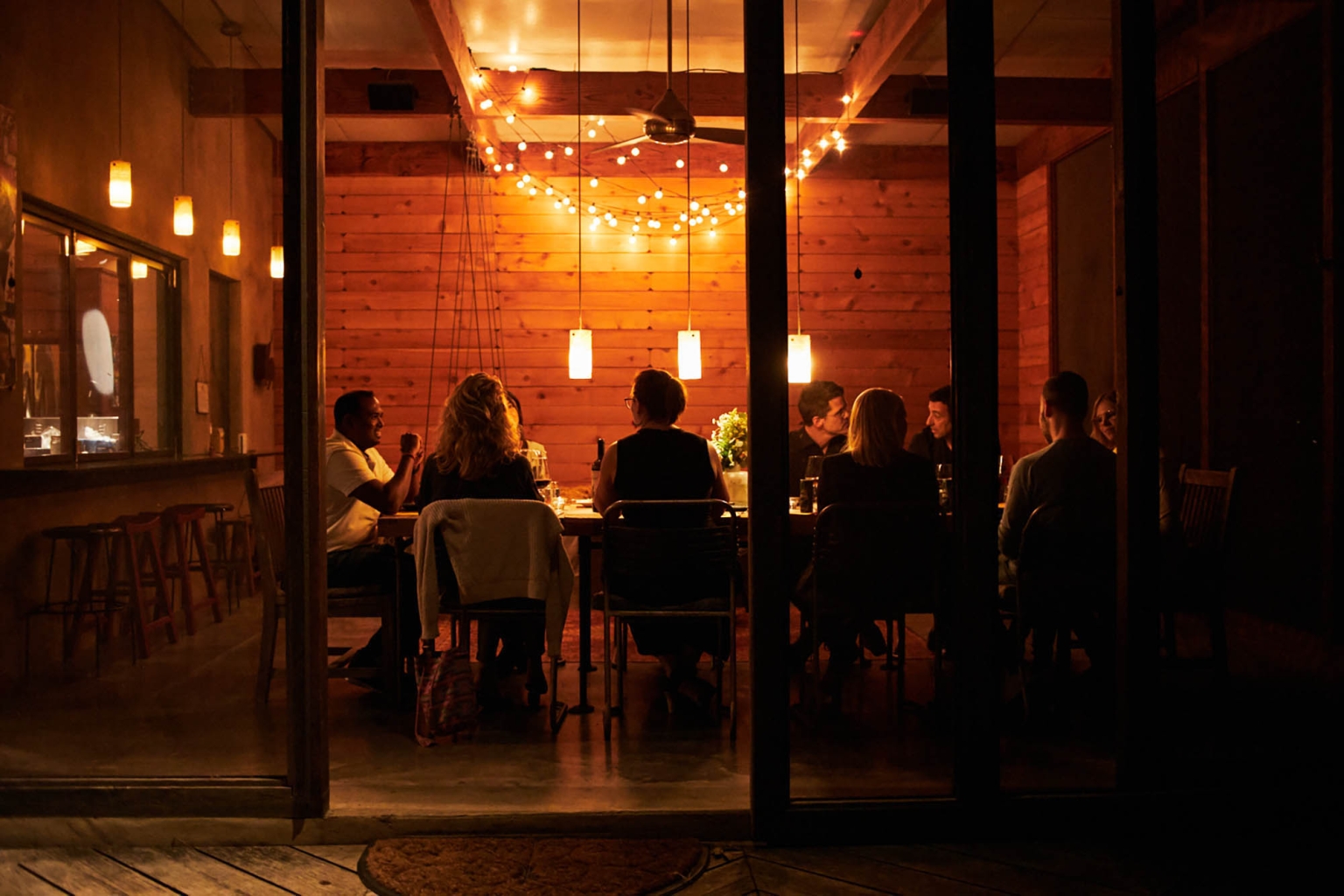 People gathered around a wooden table indoors at night, illuminated by string lights and pendant lights, creating a warm ambiance.