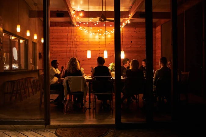 People gathered around a wooden table indoors at night, illuminated by string lights and pendant lights, creating a warm ambiance.