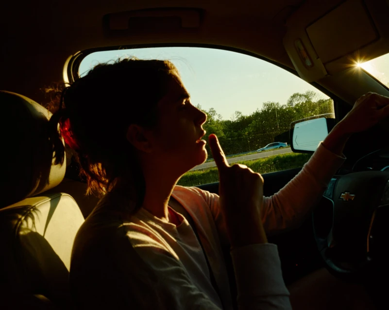 Woman driving a car on a sunny day, holding one finger up. Trees are visible through the car window, sun glare from the horizon.