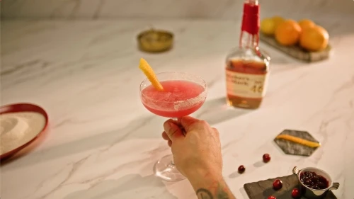 A pink cocktail in a coupe glass with a lemon twist garnish is held by a hand, set against a marble countertop with various drink ingredients.