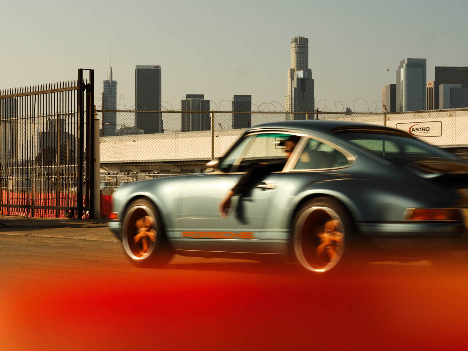 Vintage blue Porsche 911 speeding through an urban landscape with downtown Los Angeles skyscrapers in the background.