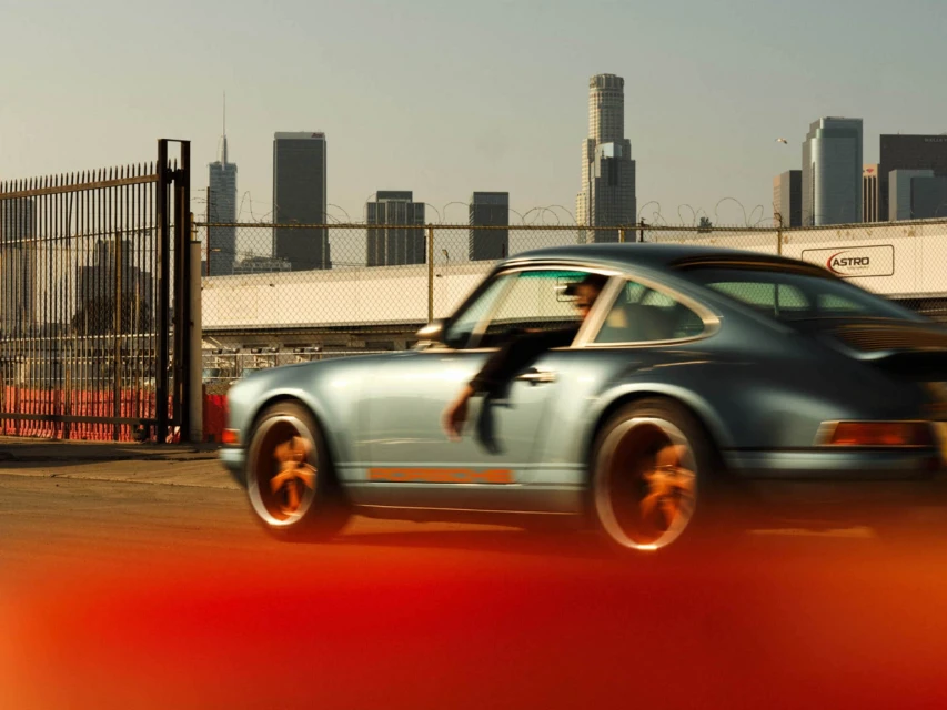 Vintage blue Porsche 911 speeding through an urban landscape with downtown Los Angeles skyscrapers in the background.