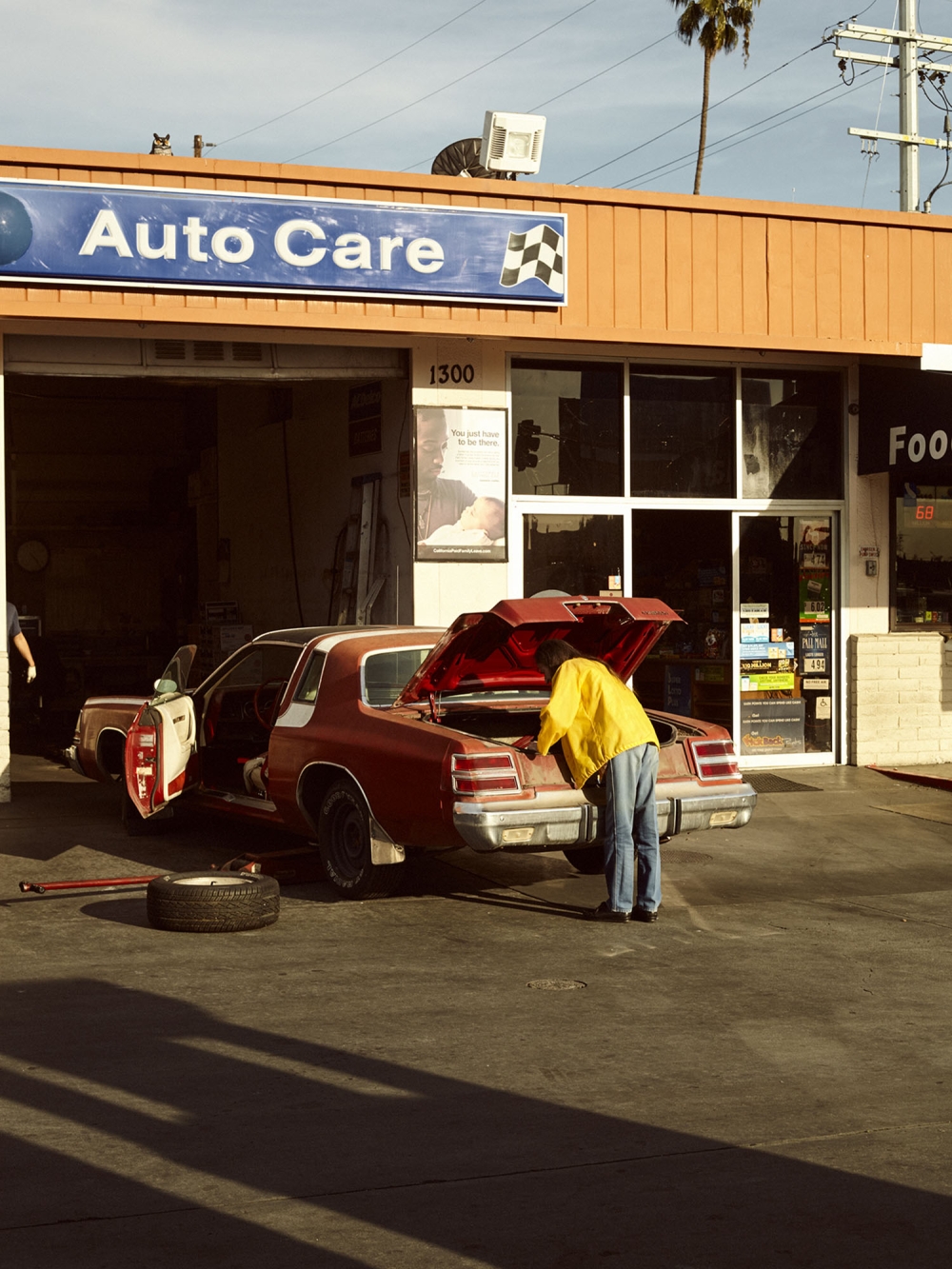 A man in a yellow jacket works on a car in front of an Auto Care garage on a sunny day with palm trees visible in the background.