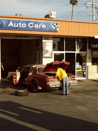 A man in a yellow jacket works on a car in front of an Auto Care garage on a sunny day with palm trees visible in the background.