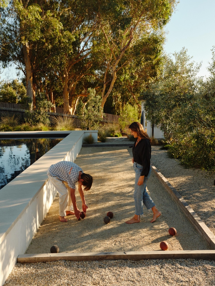 Woman and a child play bocce ball on a gravel court next to a pool, surrounded by trees, under a blue sky, outdoors in the daytime.