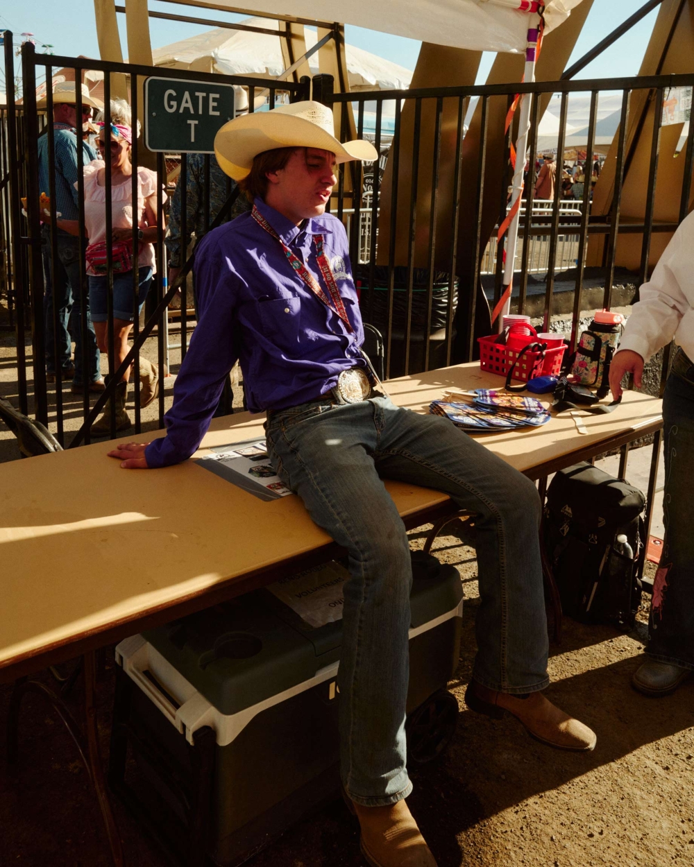 Young man wearing a cowboy hat and a purple shirt sits on a table at an outdoor event in the daytime. People in background.