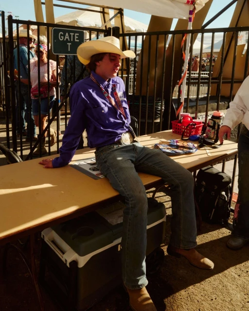 Young man wearing a cowboy hat and a purple shirt sits on a table at an outdoor event in the daytime. People in background.