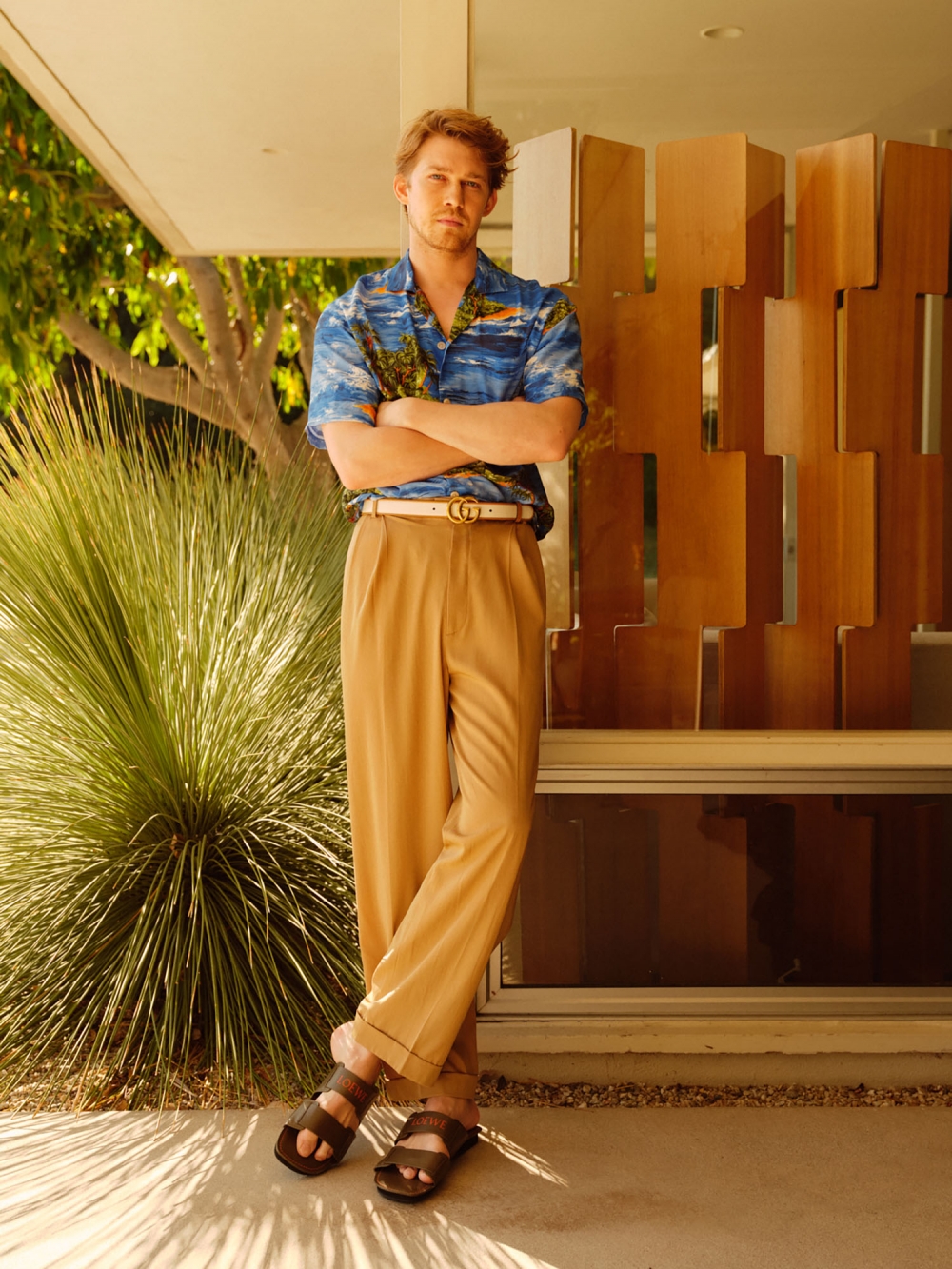 Man posing outdoors in a tropical-print shirt and tan pants, with arms crossed, standing near a building and greenery on a sunny day.
