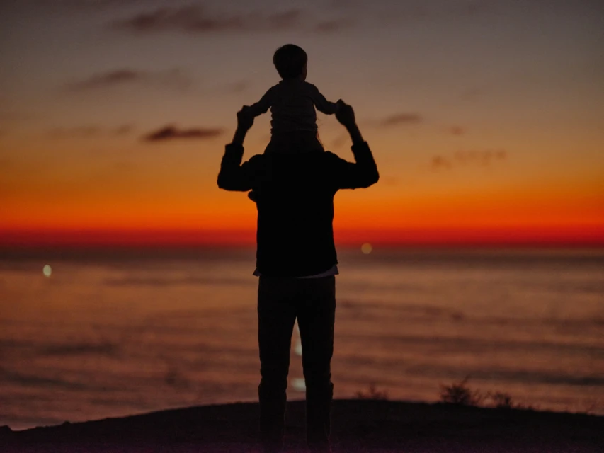 Silhouette of parent carrying child on shoulders at sunset near the ocean, creating a peaceful family moment with vibrant colors.