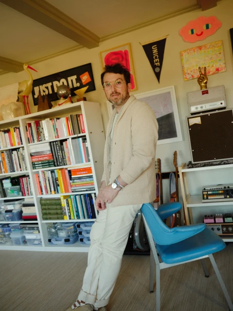 Adult man in glasses leans against blue chair in a room with a bookshelf, posters, and decor, bathed in natural light.