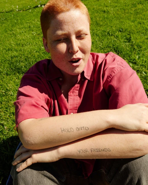 Young adult with red hair sits in sunlit green grass, wearing a red shirt and grey pants, with 'Hold onto your friends' tattooed on forearms.
