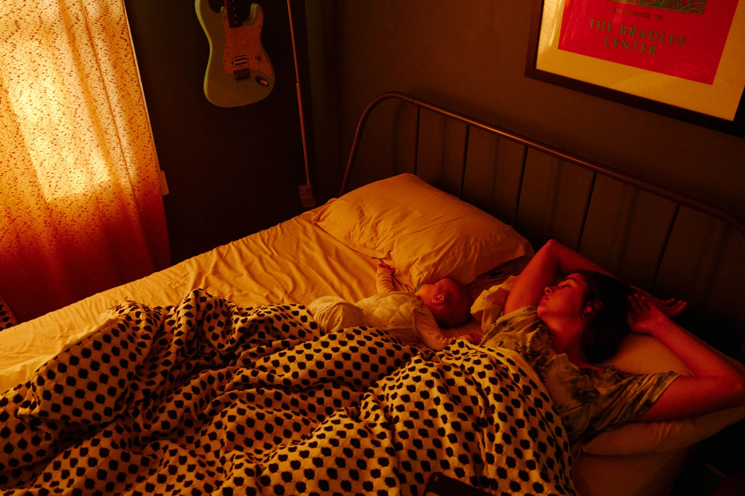 An adult female sleeps in a bed next to an infant in a warm-toned room, with a blanket, headboard, guitar, and framed art visible.