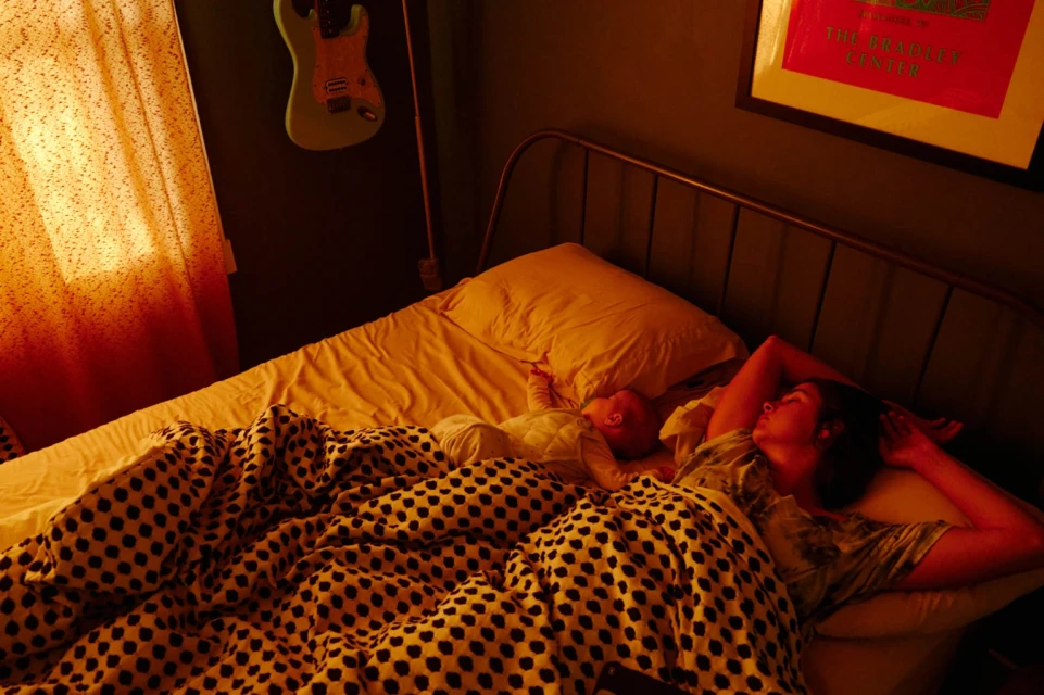 An adult female sleeps in a bed next to an infant in a warm-toned room, with a blanket, headboard, guitar, and framed art visible.