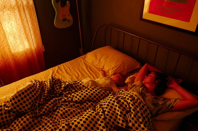 An adult female sleeps in a bed next to an infant in a warm-toned room, with a blanket, headboard, guitar, and framed art visible.