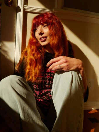Portrait of a woman with long bright red hair wearing a dark t-shirt and light jeans sitting in direct sunlight.