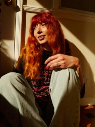 Portrait of a woman with long bright red hair wearing a dark t-shirt and light jeans sitting in direct sunlight.