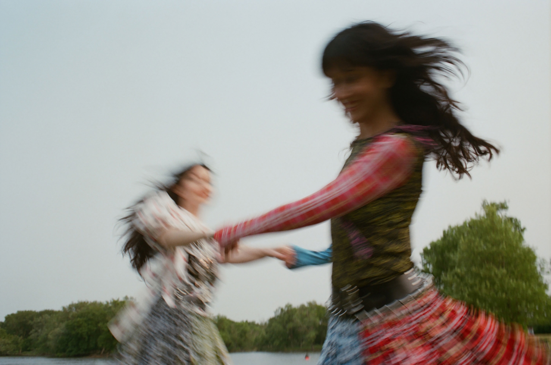 Two women in patterned dresses are holding hands, dancing, and blurred in motion against a background of trees and a cloudy sky.