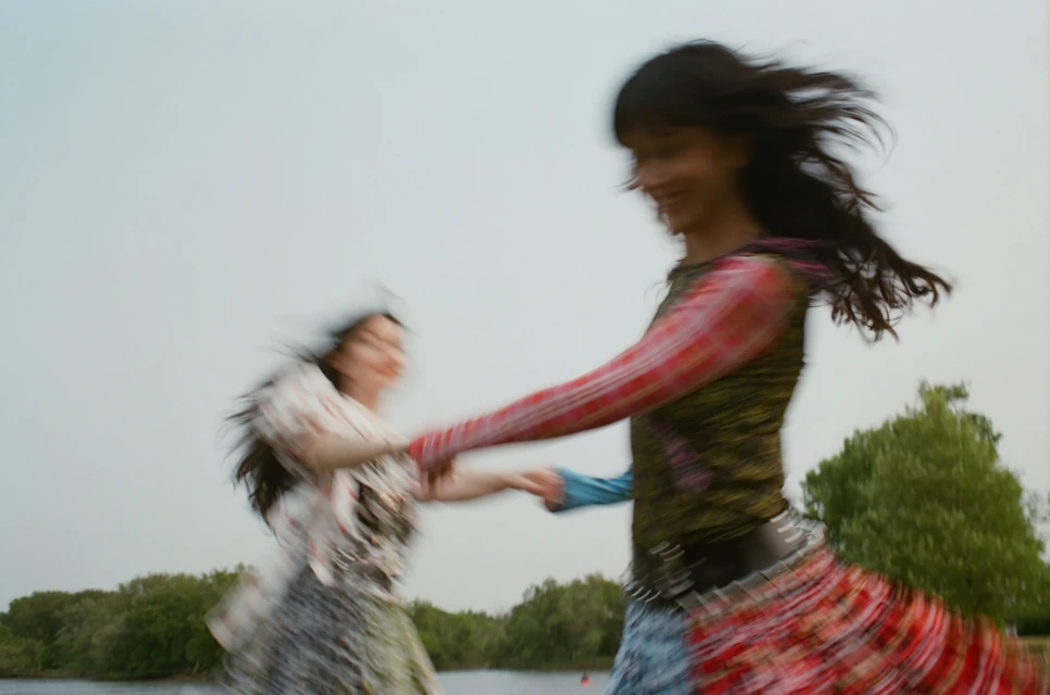 Two women in patterned dresses are holding hands, dancing, and blurred in motion against a background of trees and a cloudy sky.