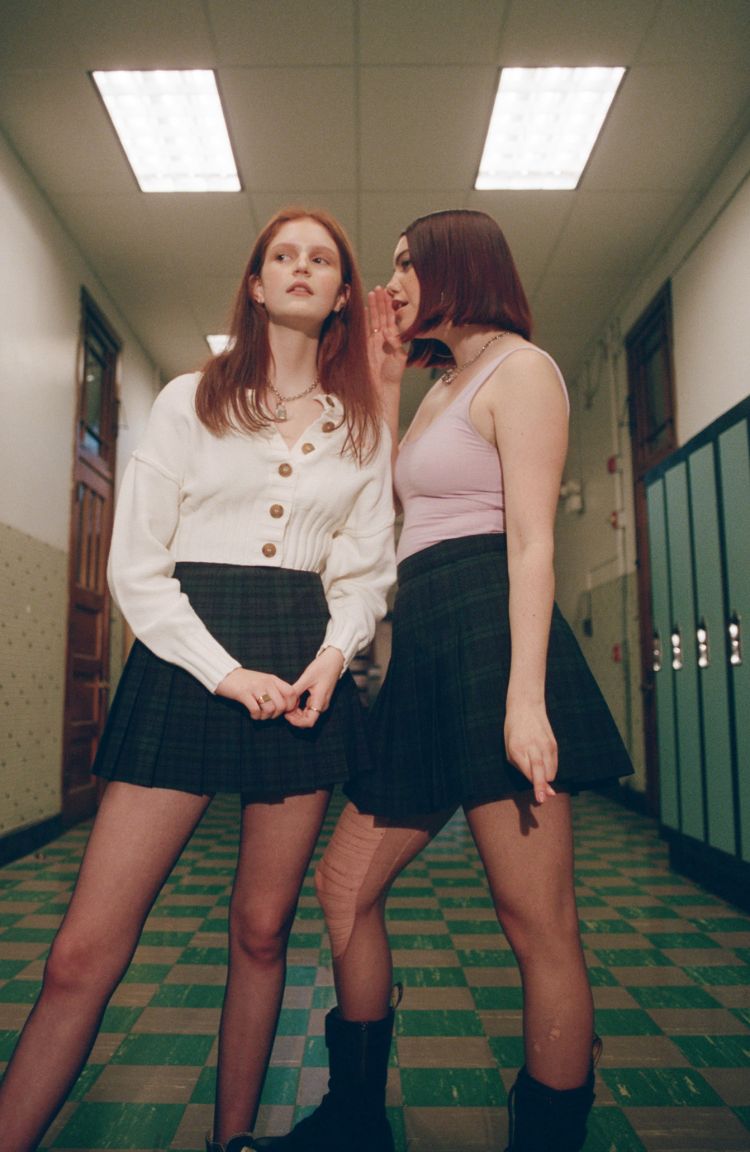 Two young women in a school hallway. One whispers into the other's ear, wearing mini skirts, cardigans, and black tights with boots.