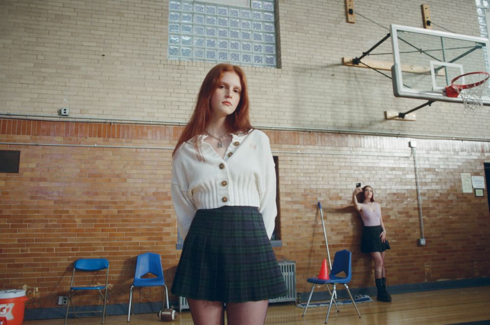 Young woman with long red hair poses in a school gymnasium wearing a white cardigan and plaid skirt, with another woman in background.