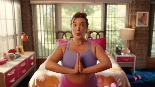 Woman in workout gear strikes a prayer pose in bedroom with orange and pink decor, sunlight streaming through the window.