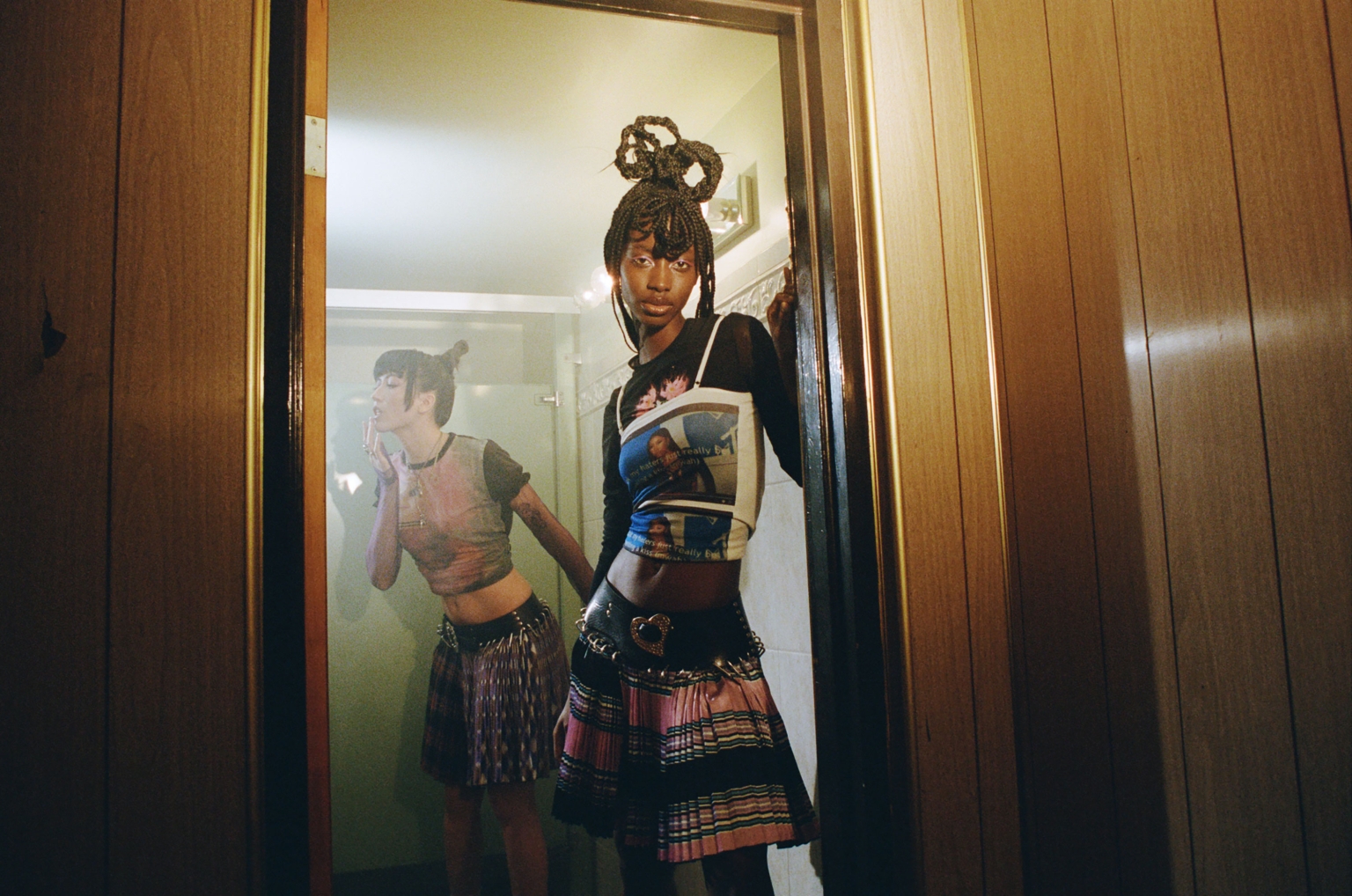 Two young women in stylish clothing pose in a bathroom; one is visible through the doorway's mirror, both with intricate braided hairstyles.
