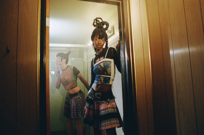 Two young women in stylish clothing pose in a bathroom; one is visible through the doorway's mirror, both with intricate braided hairstyles.