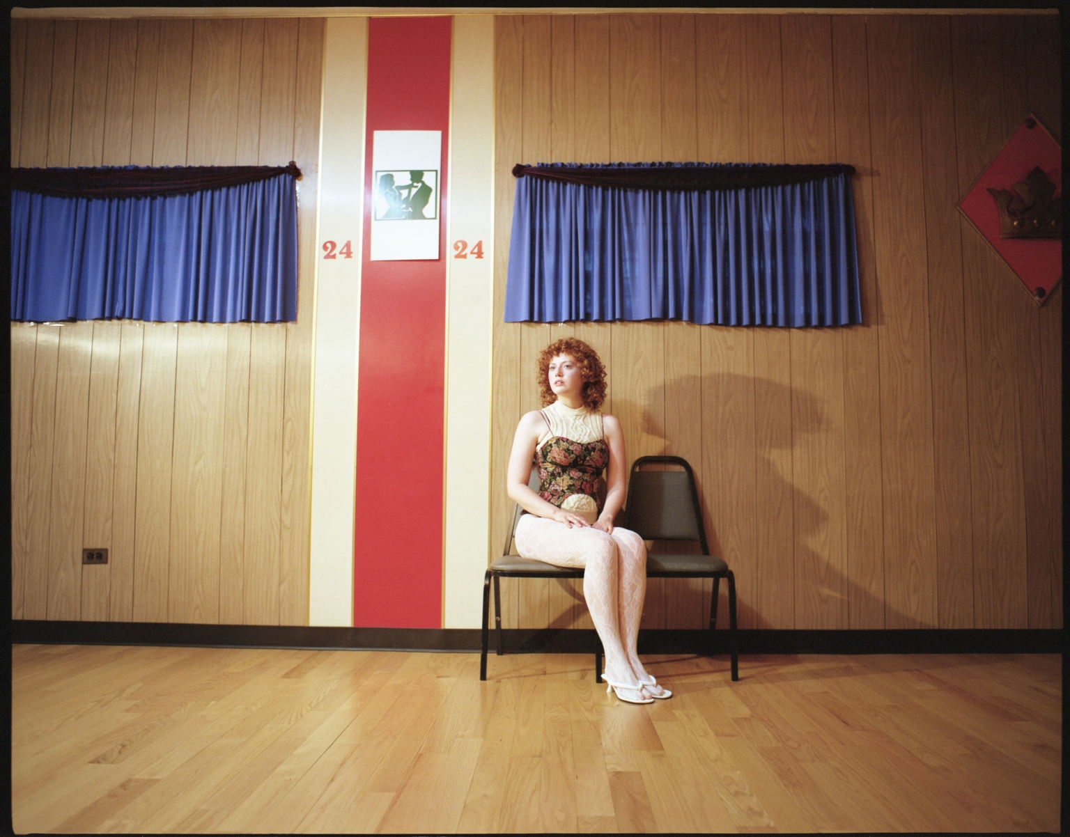 A woman with red curly hair sits on a black chair in an indoor setting with paneled walls and blue curtain accents near the wall.