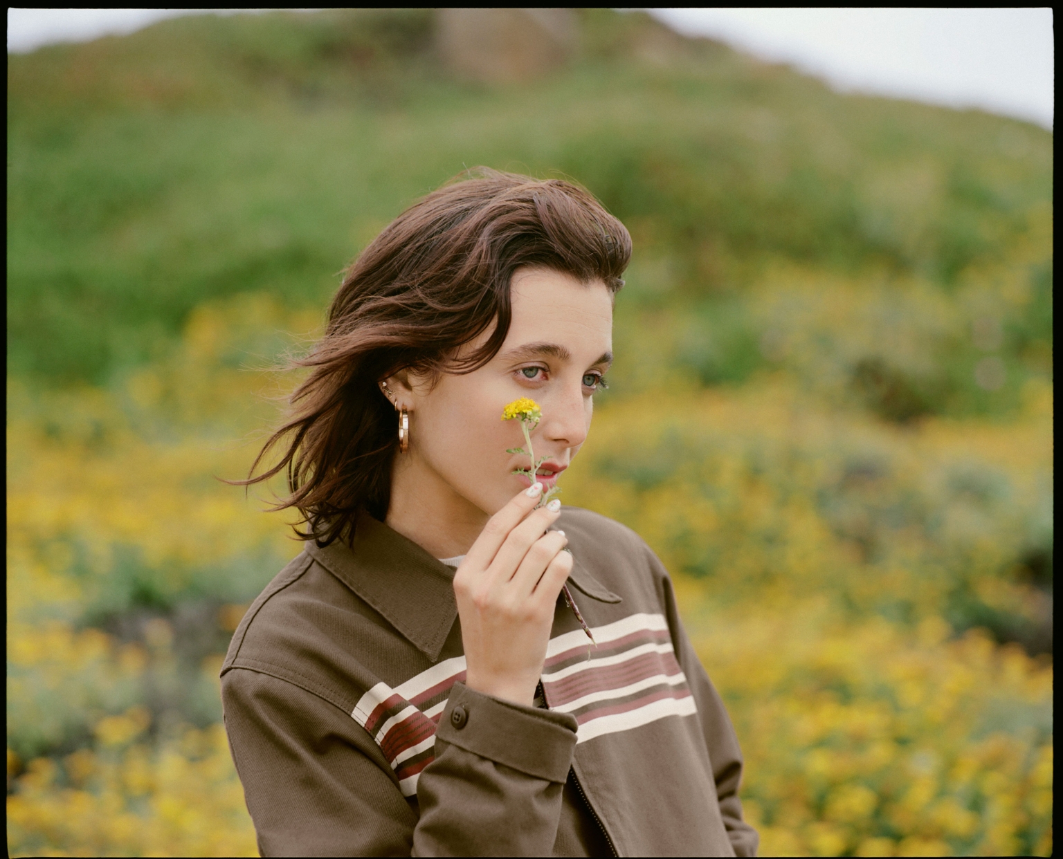 Young woman with short brown hair holds a yellow dandelion in a field of yellow flowers, wearing a brown jacket. Outdoor portrait.