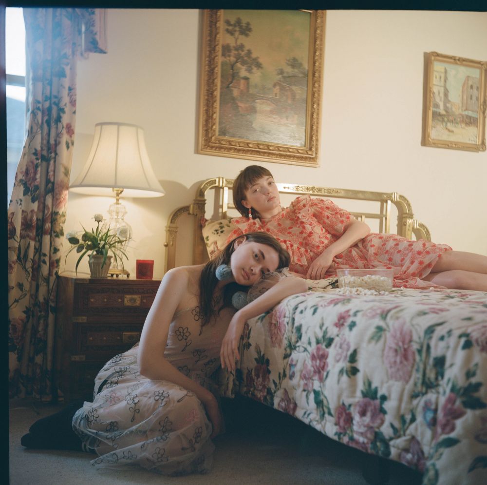 Two young women pose on a floral bed inside a brightly lit room with floral curtains and gold-framed landscape painting on the wall.