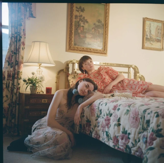 Two young women pose on a floral bed inside a brightly lit room with floral curtains and gold-framed landscape painting on the wall.