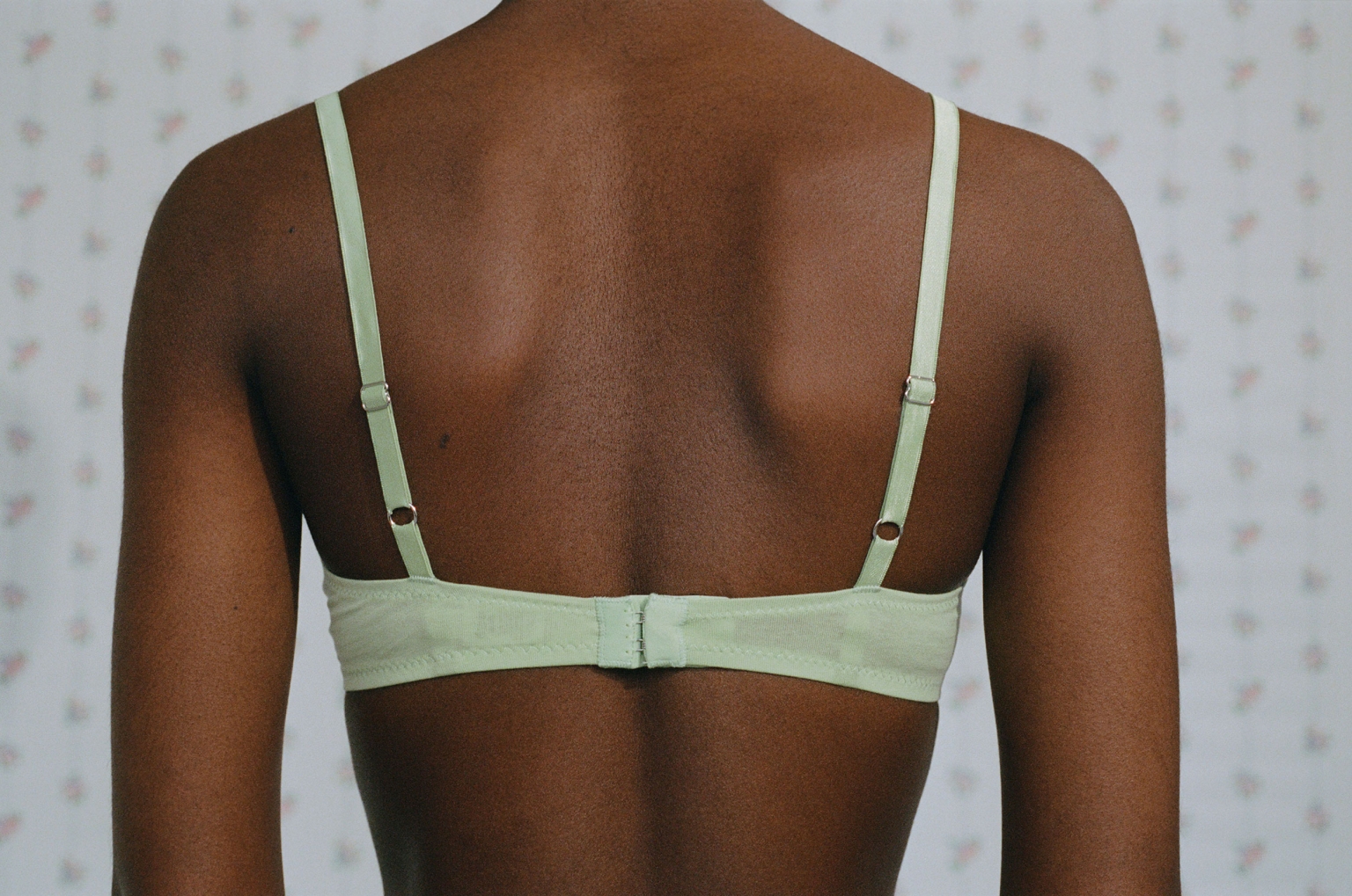 Close-up back view of a dark-skinned adult female in a light green bra against a light background with small pink floral pattern.