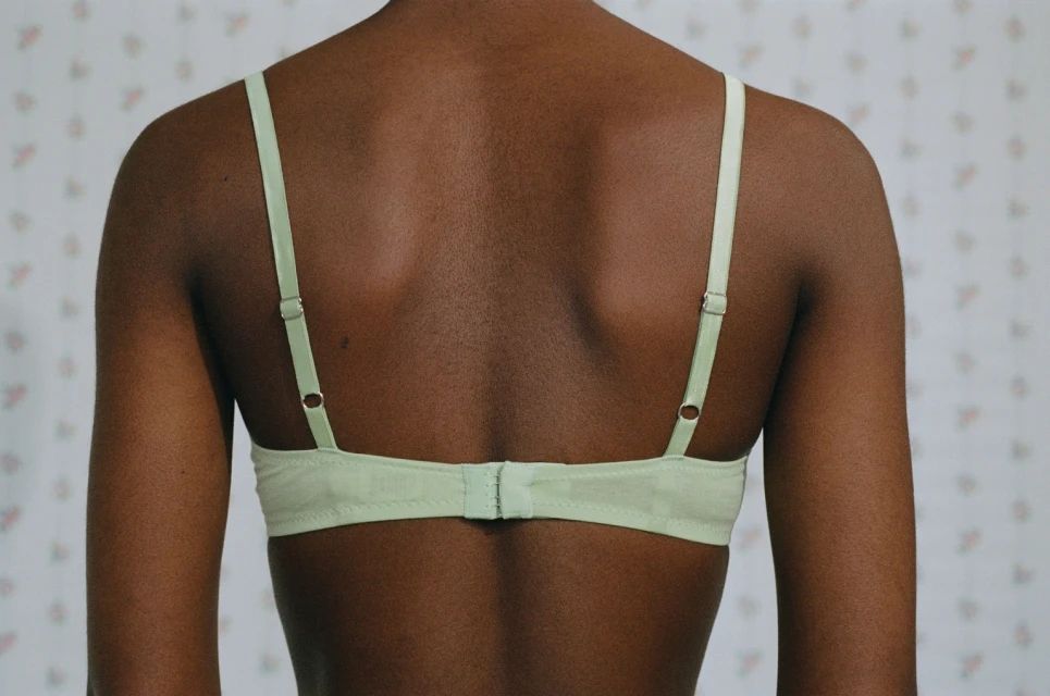Close-up back view of a dark-skinned adult female in a light green bra against a light background with small pink floral pattern.