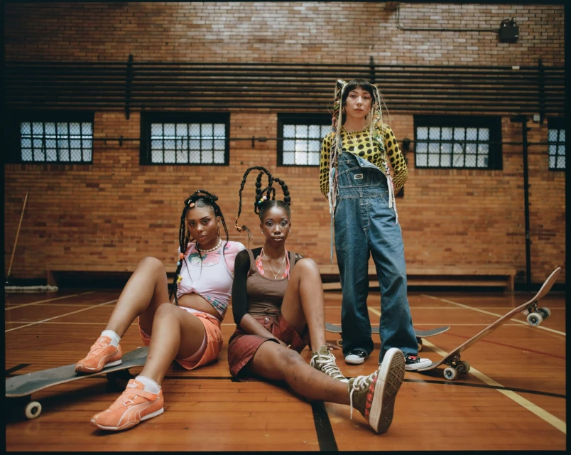 Three young women pose in a gymnasium wearing trendy clothing and sneakers, skateboards nearby, showcasing a modern urban fashion shoot.