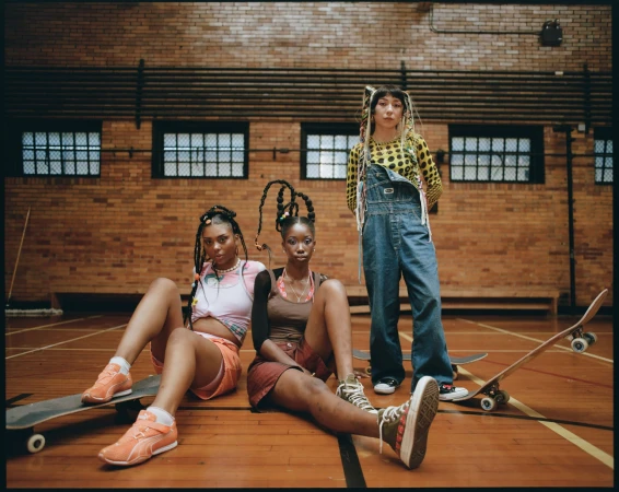 Three young women pose in a gymnasium wearing trendy clothing and sneakers, skateboards nearby, showcasing a modern urban fashion shoot.