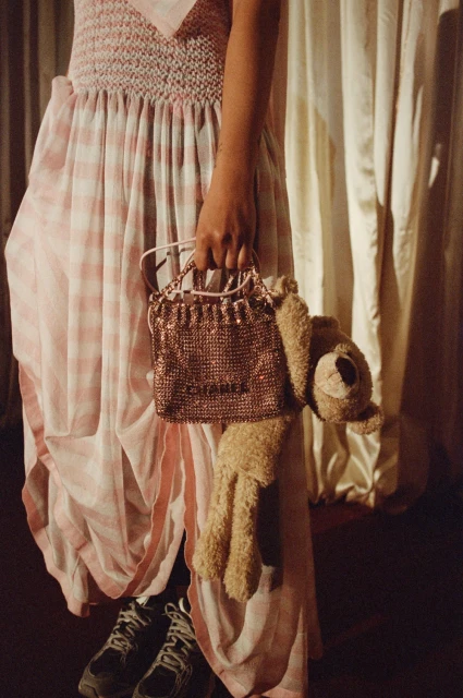 Woman in checkered dress holds sparkly purse with bear. Casual fashion with plush toy, sneakers, and draped fabric backdrop.