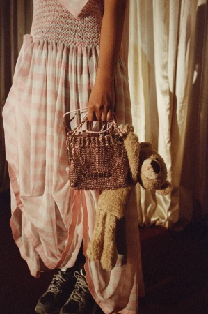 Woman in checkered dress holds sparkly purse with bear. Casual fashion with plush toy, sneakers, and draped fabric backdrop.