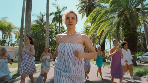 A group of women are running outside wearing towels, palm trees line the street, and sunlight creates a bright, summery atmosphere.