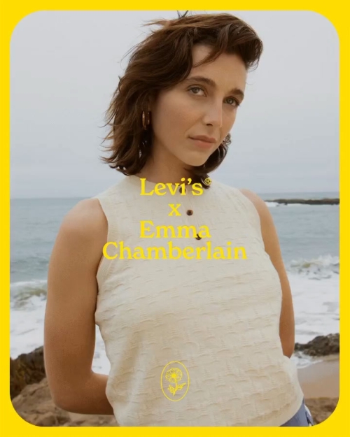 Emma Chamberlain poses in front of a beach and ocean, wearing a cream top, earrings, and a neutral makeup palette.