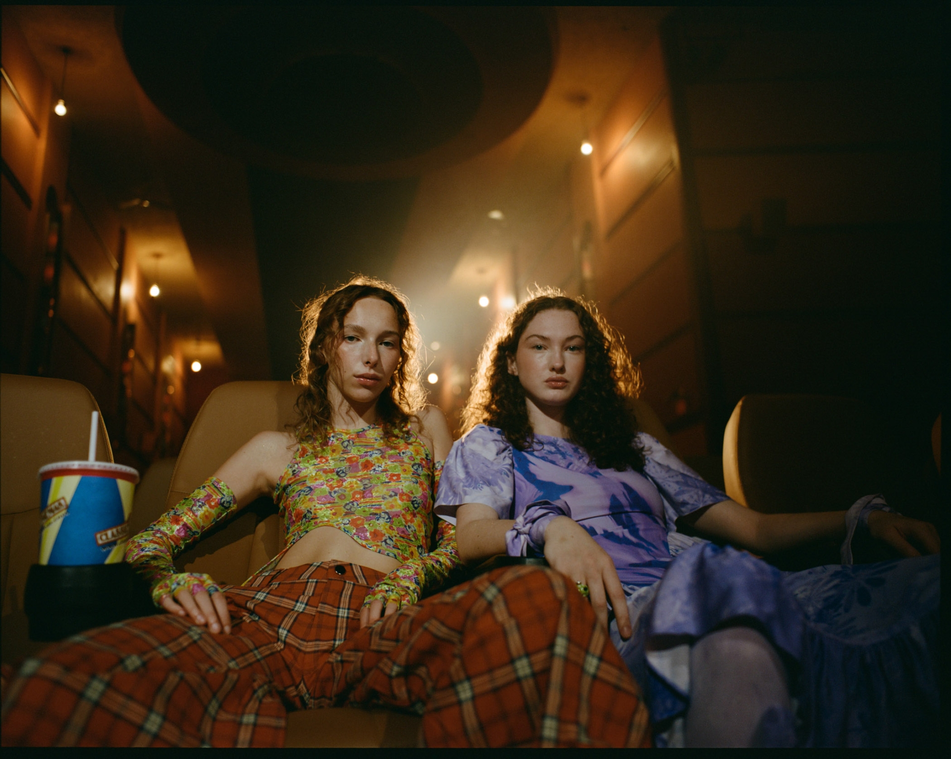 Two women sit in a movie theater wearing retro-style outfits with a drink. Warm lighting and a vintage aesthetic create a cozy scene.