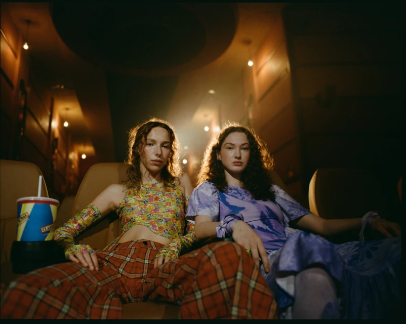 Two women sit in a movie theater wearing retro-style outfits with a drink. Warm lighting and a vintage aesthetic create a cozy scene.