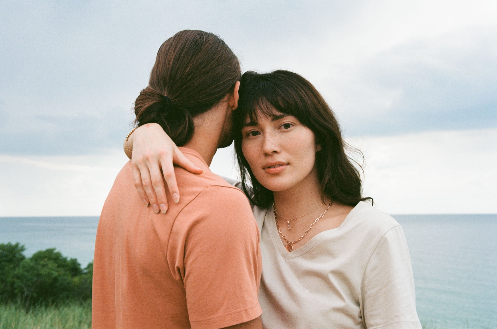A young couple embrace near the ocean on an overcast day. The brunette woman looks at the camera with a serious expression.