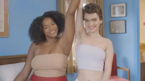 Two smiling women pose indoors in a bedroom. They are wearing tube tops and have their arms raised to show their armpits.