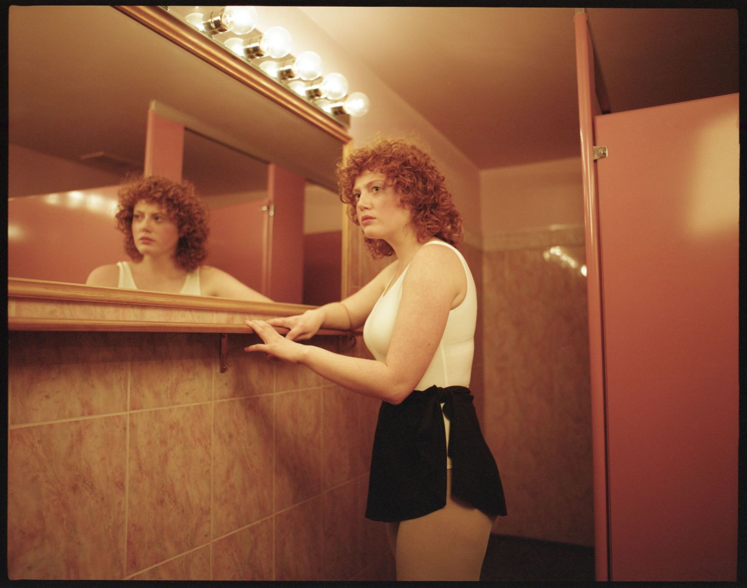 A woman with curly red hair poses in a bathroom, reflected in a mirror with a light fixture above, wearing a white top and black skirt.