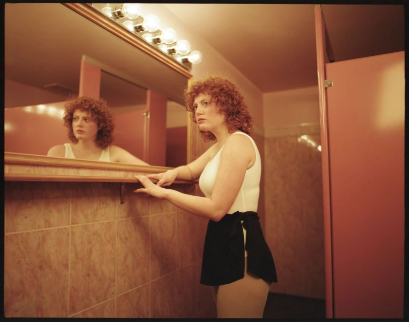 A woman with curly red hair poses in a bathroom, reflected in a mirror with a light fixture above, wearing a white top and black skirt.