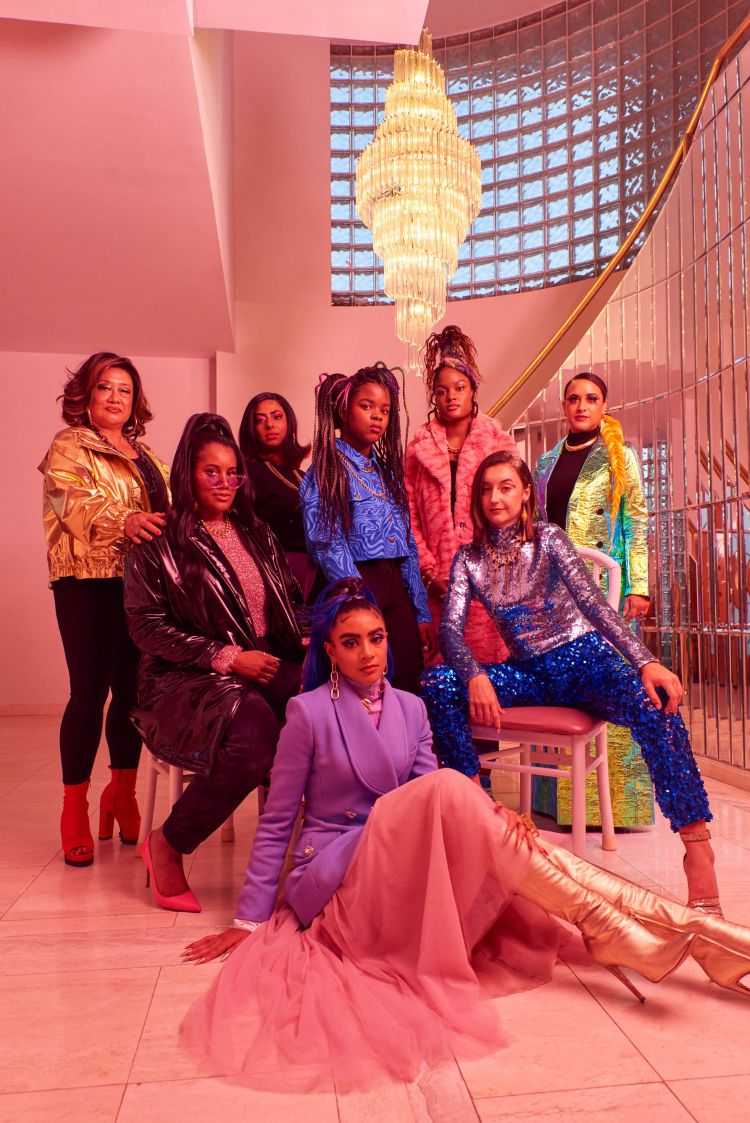 Group portrait of eight fashionable women in a pink-toned interior featuring a crystal chandelier and architectural details.