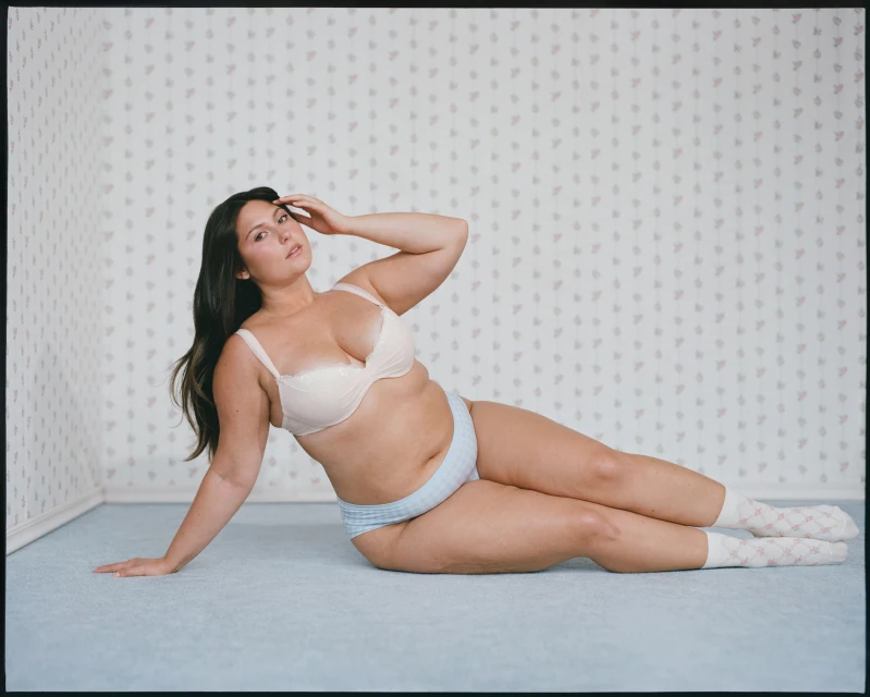 Plus-size woman poses in bra and underwear against a patterned wall. Indoor studio shot with soft lighting, showing body positivity.