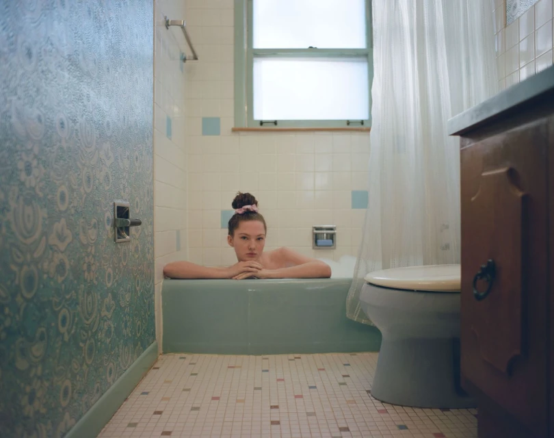 Young woman relaxes in a turquoise bathtub, eyes forward, set in a retro-styled bathroom with patterned wallpaper and vintage fixtures.